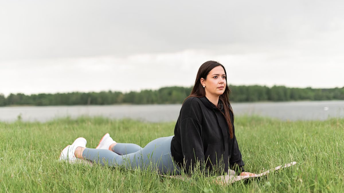 Woman lying on a yoga mat in a grassy field by a lake, promoting relaxation and wellness.
