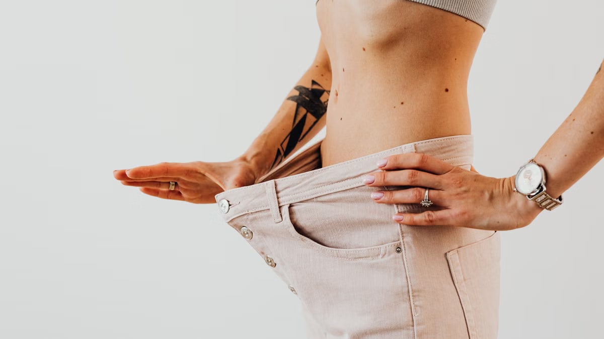 Close-up of a woman in oversized pants showcasing weight loss progress. Studio shot with neutral background.